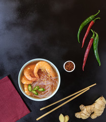 Asian soup with shrimps, rice noodles, ginger and hot pepper on a black background. Bamboo chopsticks near the plate. Copy space.