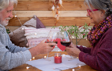 A smiling senior couple show two red heart shapes. sitting outdoor in a romantic corner made by wood. Yellow lights. Two red wineglasses to celebrate a positive moment