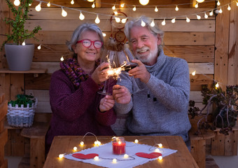 A smiling senior couple with sparkles celebrate the incoming new year in a romantic corner made by wood. Yellow lights. Two red wineglasses to wish a happy 2020