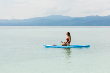 Woman in red swimsuit sitting on sup board and looking on horizon view with blue mountain silhouettes. Tropical travel and water activity concept. Empty space and water background.