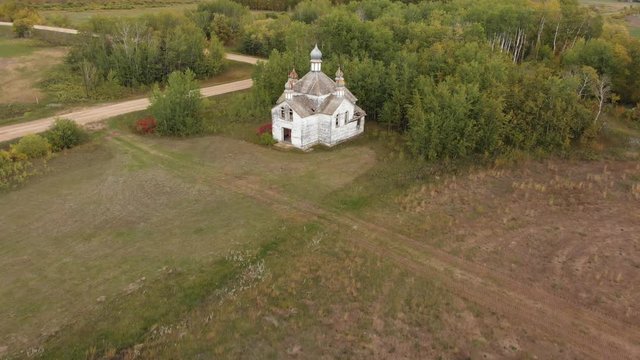High flying rone moving towards an abandoned and dilapidated white church surrounded with trees and farm fields.