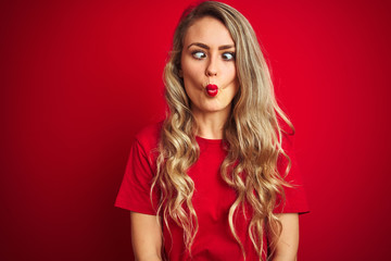Fototapeta premium Young beautiful woman wearing basic t-shirt standing over red isolated background making fish face with lips, crazy and comical gesture. Funny expression.