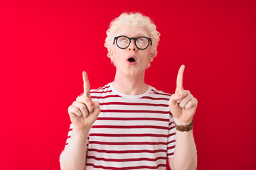 Young albino blond man wearing striped t-shirt and glasses over isolated red background amazed and...
