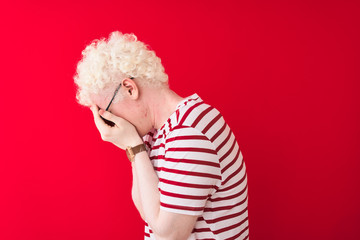 Young albino blond man wearing striped t-shirt and glasses over isolated red background with sad expression covering face with hands while crying. Depression concept.