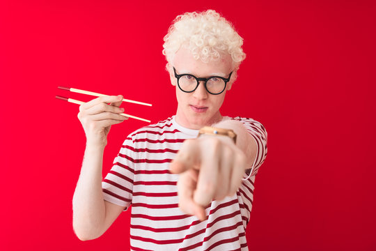 Young albino blond man holding chopsticks standing over isolated white background pointing with finger to the camera and to you, hand sign, positive and confident gesture from the front