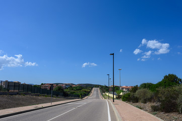 Fototapeta premium Road goes into the distance against a blue sky with clouds. On the sides trees and a small village