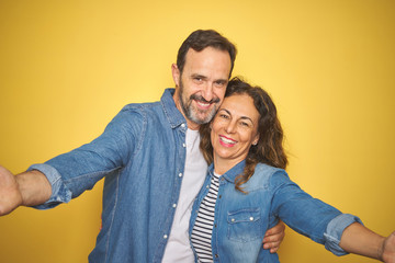 Beautiful middle age couple together wearing denim shirt over isolated yellow background looking at the camera smiling with open arms for hug. Cheerful expression embracing happiness.