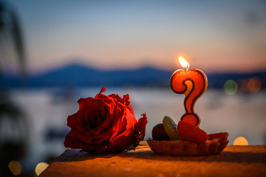 Burning candle in the form of a question mark and a rose close up selective focus on the sunset against the sea and palm trees