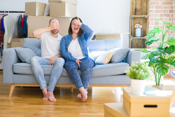 Young couple sitting on the sofa arround cardboard boxes moving to a new house smiling and laughing with hand on face covering eyes for surprise. Blind concept.