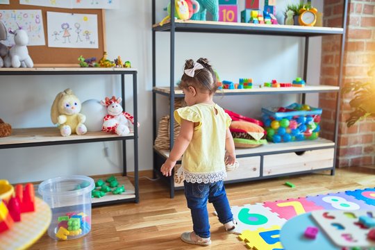 Beautiful toddler standing looking at shelving with dolls at kindergarten