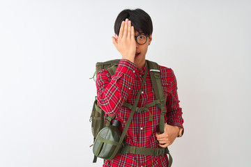 Chinese hiker man wearing backpack canteen glasses over isolated white background covering one eye with hand, confident smile on face and surprise emotion.