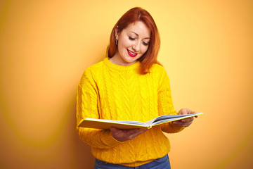 Young redhead student woman reading book standing over yellow isolated background with a happy face...