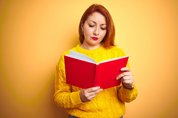 Young redhead teacher woman reading red book over yellow isolated background with a confident...