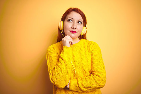 Young redhead woman listening to music using headphones over yellow isolated background with hand on chin thinking about question, pensive expression. Smiling with thoughtful face. Doubt concept.