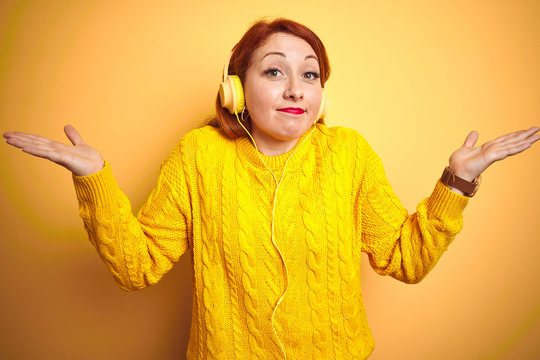 Young redhead woman listening to music using headphones over yellow isolated background clueless and confused expression with arms and hands raised. Doubt concept.