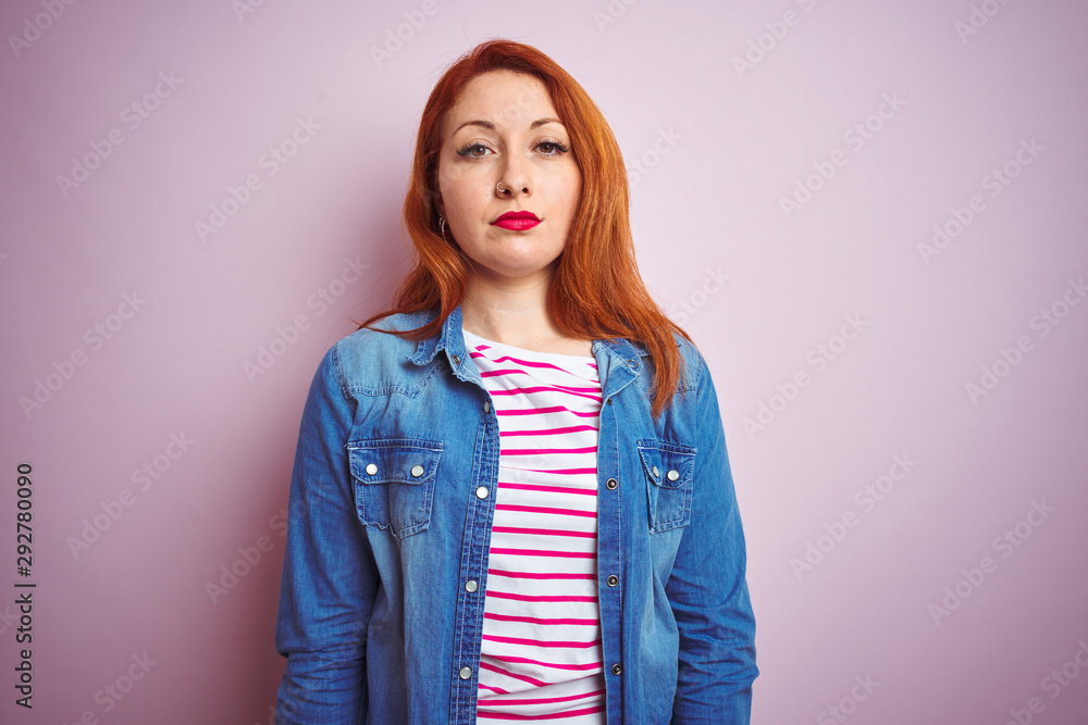 Wall mural Beautiful redhead woman wearing denim shirt and striped t-shirt over isolated pink background Relaxed with serious expression on face. Simple and natural looking at the camera.