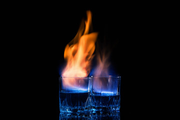 Close up, macro. Two glasses with cocktails, on the surface of which a blue-orange flame quivers. Black background. Copy space. - obrazy, fototapety, plakaty