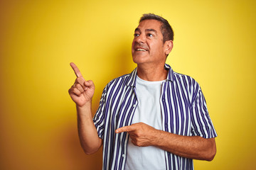 Handsome middle age man wearing striped shirt standing over isolated yellow background smiling and looking at the camera pointing with two hands and fingers to the side.