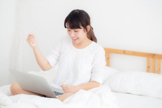 Beautiful of portrait asian young woman sitting relax and leisure with laptop computer internet online on bed in bedroom, cheerful of asia girl with gesture glad and success, lifestyle concept.