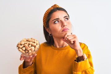 Young beautiful woman holding bowl with peanuts standing over isolated white background serious...