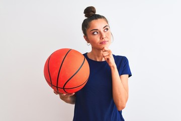 Young beautiful sportswoman holding basketball ball over isolated white background serious face thinking about question, very confused idea