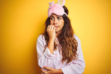 Young woman wearing pajama and sleep mask standing over yellow isolated background looking stressed and nervous with hands on mouth biting nails. Anxiety problem.