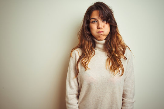 Young beautiful woman wearing winter sweater standing over white isolated background puffing cheeks with funny face. Mouth inflated with air, crazy expression.