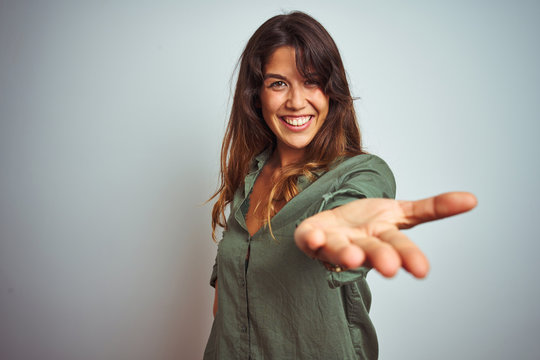 Young beautiful woman wearing green shirt standing over grey isolated background smiling cheerful offering palm hand giving assistance and acceptance.