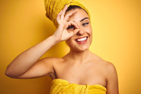 Young beautiful woman wearing towel after shower over isolated yellow background with happy face smiling doing ok sign with hand on eye looking through fingers