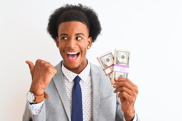 Young african american businessman holding dollars standing over isolated white background pointing and showing with thumb up to the side with happy face smiling