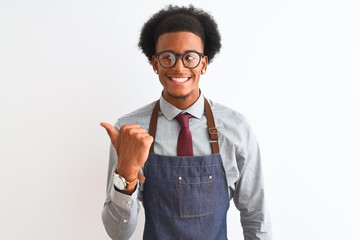 Young african american shopkeeper man wearing apron glasses over isolated white background smiling with happy face looking and pointing to the side with thumb up.