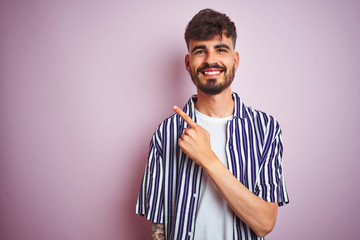 Young man with tattoo wearing striped shirt standing over isolated pink background cheerful with a smile of face pointing with hand and finger up to the side with happy and natural expression on face