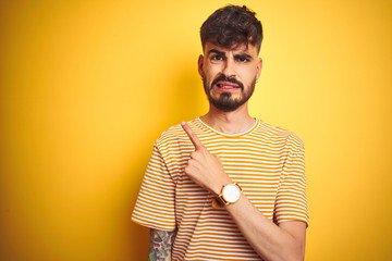 Young man with tattoo wearing striped t-shirt standing over isolated yellow background Pointing...