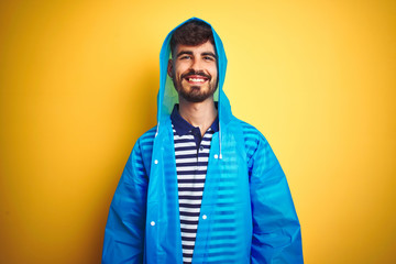 Young handsome man wearing rain coat with hood standing over isolated yellow background with a happy and cool smile on face. Lucky person.