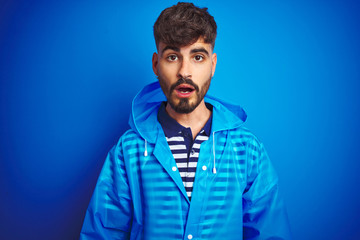 Young handsome man wearing rain coat standing over isolated blue background afraid and shocked with surprise expression, fear and excited face.