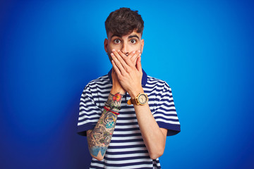 Young man with tattoo wearing striped polo standing over isolated blue background shocked covering mouth with hands for mistake. Secret concept.