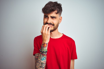 Young man with tattoo wearing red t-shirt standing over isolated white background looking stressed and nervous with hands on mouth biting nails. Anxiety problem.
