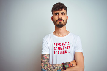 Young man with tattoo wearing fanny t-shirt standing over isolated white background skeptic and...