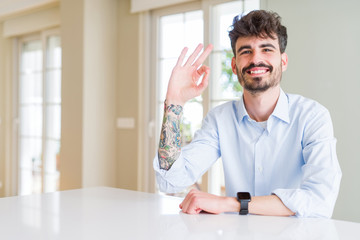 Young businesss man sitting on white table smiling positive doing ok sign with hand and fingers. Successful expression.