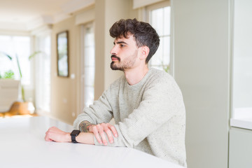 Young man wearing casual sweater sitting on white table looking to side, relax profile pose with natural face with confident smile.