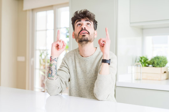 Young man wearing casual sweater sitting on white table amazed and surprised looking up and pointing with fingers and raised arms.