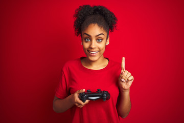 African american gamer woman playing video game using joystick over isolated red background surprised with an idea or question pointing finger with happy face, number one © Krakenimages.com