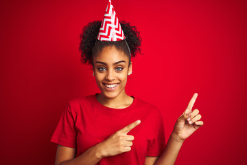 Young afro american woman wearing birthday hat over isolated red background smiling and looking at...
