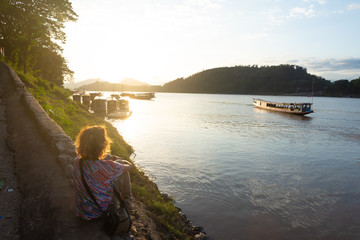 Woman looking at boats on Mekong River at Luang Prabang Laos, sunset dramatic sky, famous travel destination backpacker in South East Asia