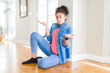 Beautiful young african american woman with afro hair sitting on the floor clueless and confused...