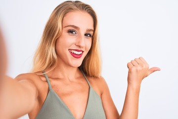 Young beautiful woman wearing t-shirt make selfie by camera over isolated white background pointing and showing with thumb up to the side with happy face smiling