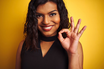 Transsexual transgender woman wearing black t-shirt over isolated yellow background doing ok sign with fingers, excellent symbol