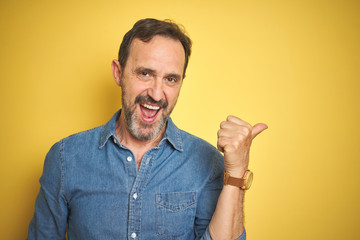 Handsome middle age senior man with grey hair over isolated yellow background smiling with happy face looking and pointing to the side with thumb up.