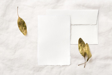 minimalist hand made paper greeting card or wedding stationery mockup with card and envelope as well as elegant green, dry autumn / fall leaves on a soft, bright, feminine fabric background   