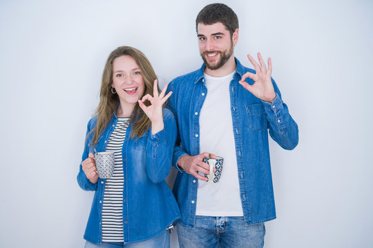 Young beautiful couple drinking a cup of coffee over white isolated background doing ok sign with fingers, excellent symbol